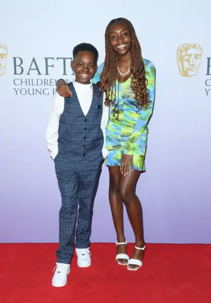 A young boy and a young girl on the red carpet. The boy is wearing a blue checked waistcoat and trousers and the girl is wearing a blue and green patterned dress.