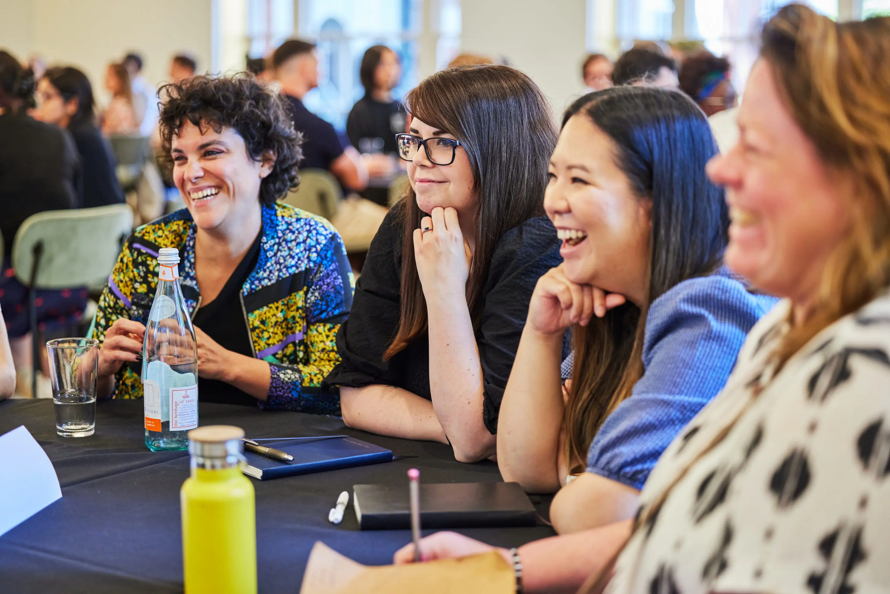 Three dark-haired women smiling and chatting. The woman on the left has short curly hair and is wearing a bright blue and yellow bomber jacket, the woman in the middle has long straight hair, glasses and is wearing a black top and the woman on the right has long straight hair and is wearing a blue short sleeved top,