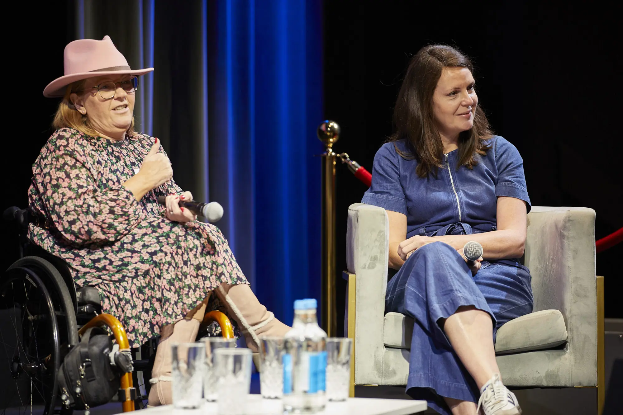 Two women sat on stage with a blue curtain behind them. The woman on the left is blonde, wearing a pink hat and sat in a gold wheelchair. The woman on the right has mid-length brown hair and is wearing a blue jumpsuit.