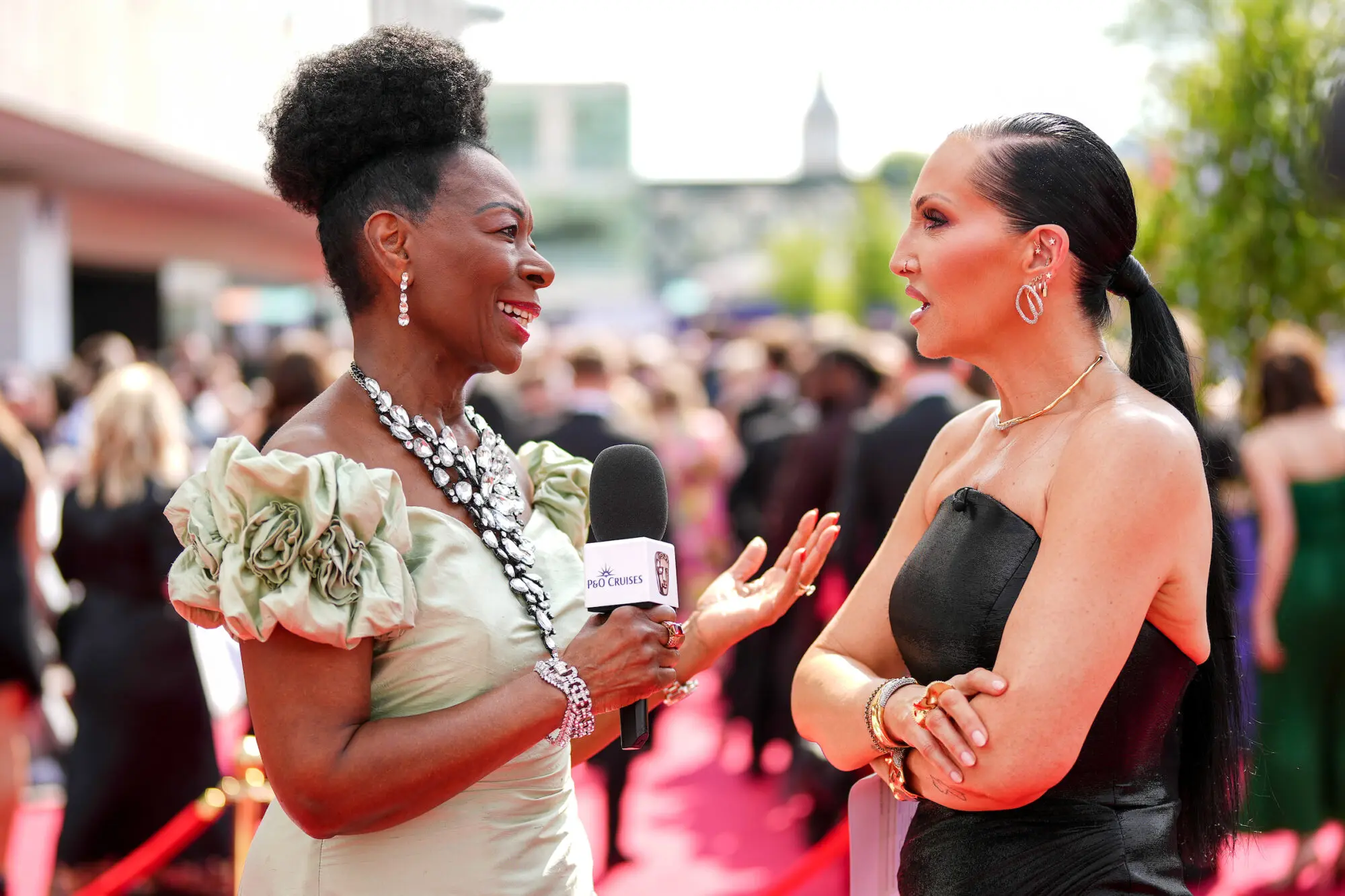A black woman in a pale green evening gown and long crystal necklace interviewing a woman in a black evening gown with dark scraped back hair.