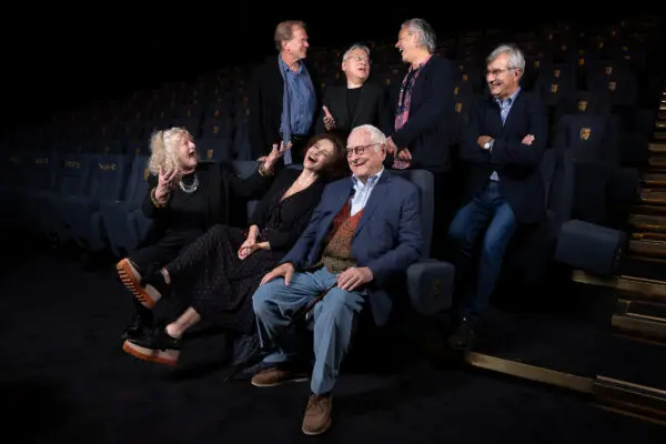 A man with white hair and glasses wearing a shirt, jumper and suit jacket is surrounded by five other people.