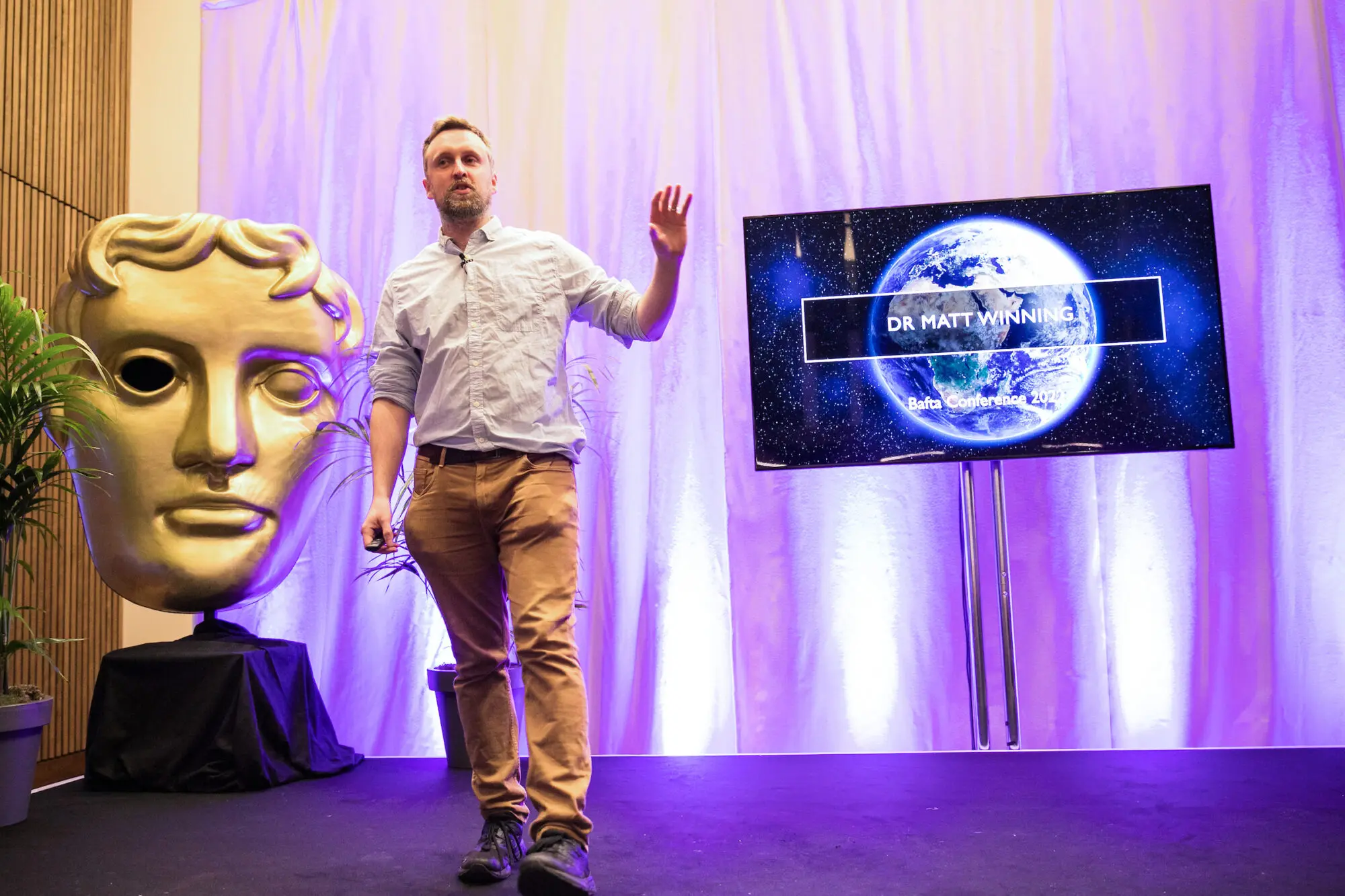 A man in a white shirt and tan trousers standing on a stage presenting next to a screen with an image of a world on it. To his left is a large gold BAFTA statue.