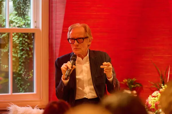 A man with grey hair and black thick rimmed glasses wearing a white shirt and black jacket. He is speaking into a microphone.