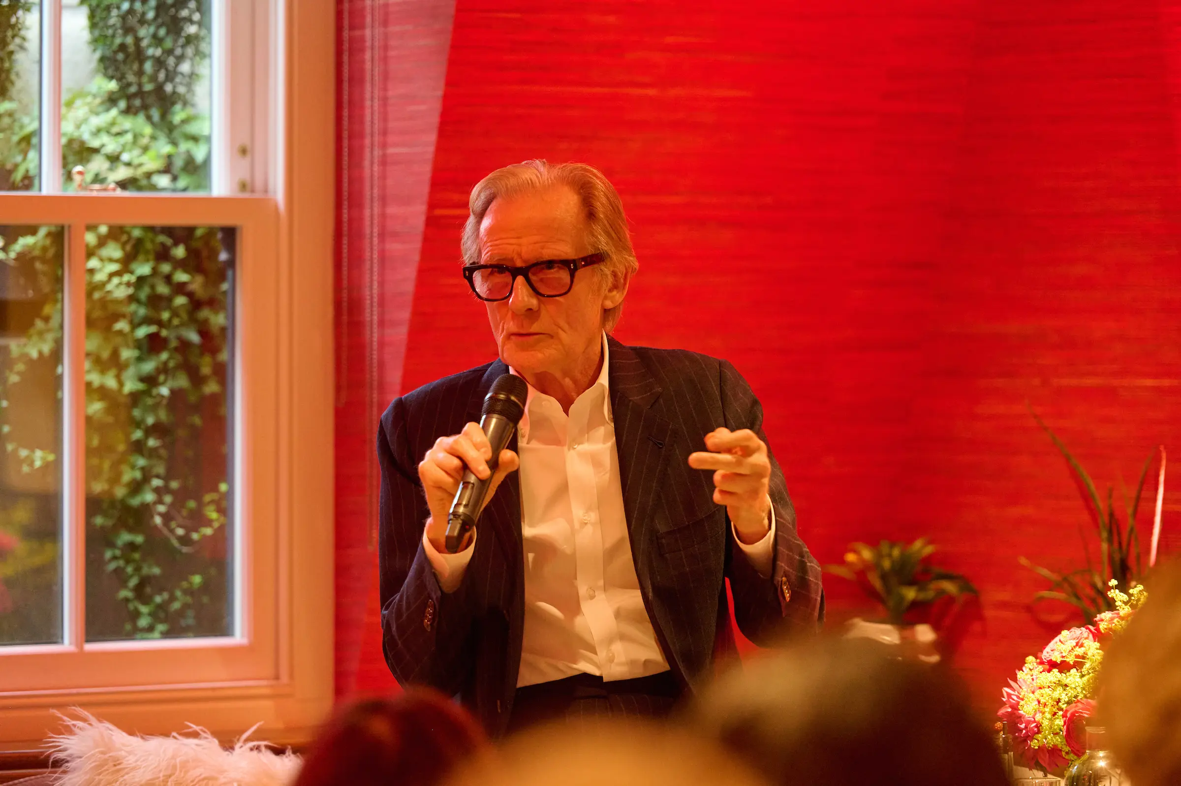 A man with grey hair and black thick rimmed glasses wearing a white shirt and black jacket. He is speaking into a microphone.
