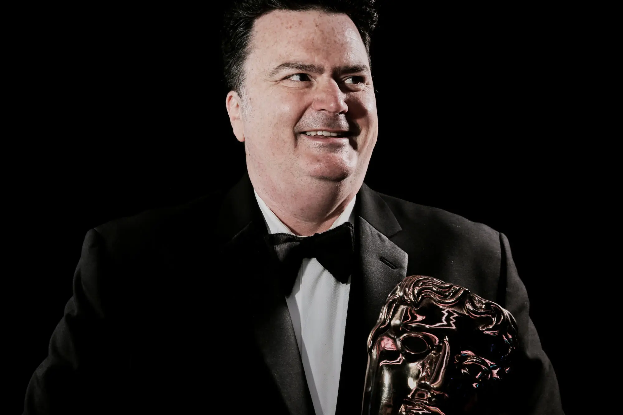 A man with short black hair wearing a black tuxedo and bow tie and holding a BAFTA.