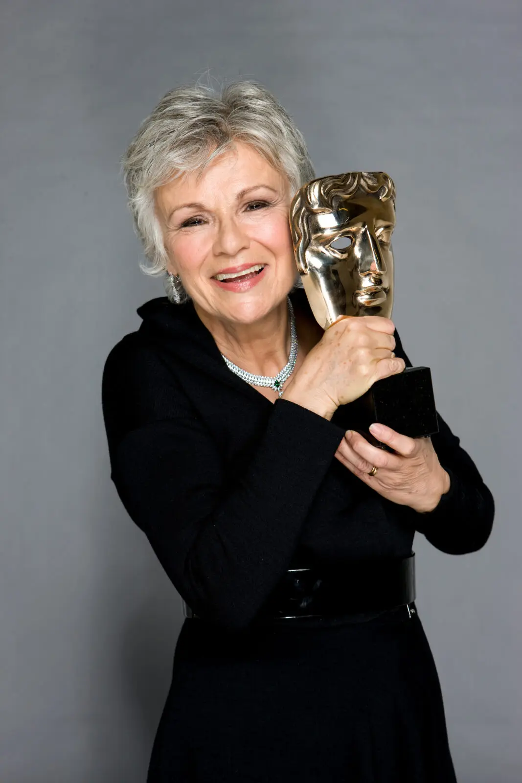 An older woman with short grey hair and wearing a black belted dress and jewelled necklace. She is holding a BAFTA mask.