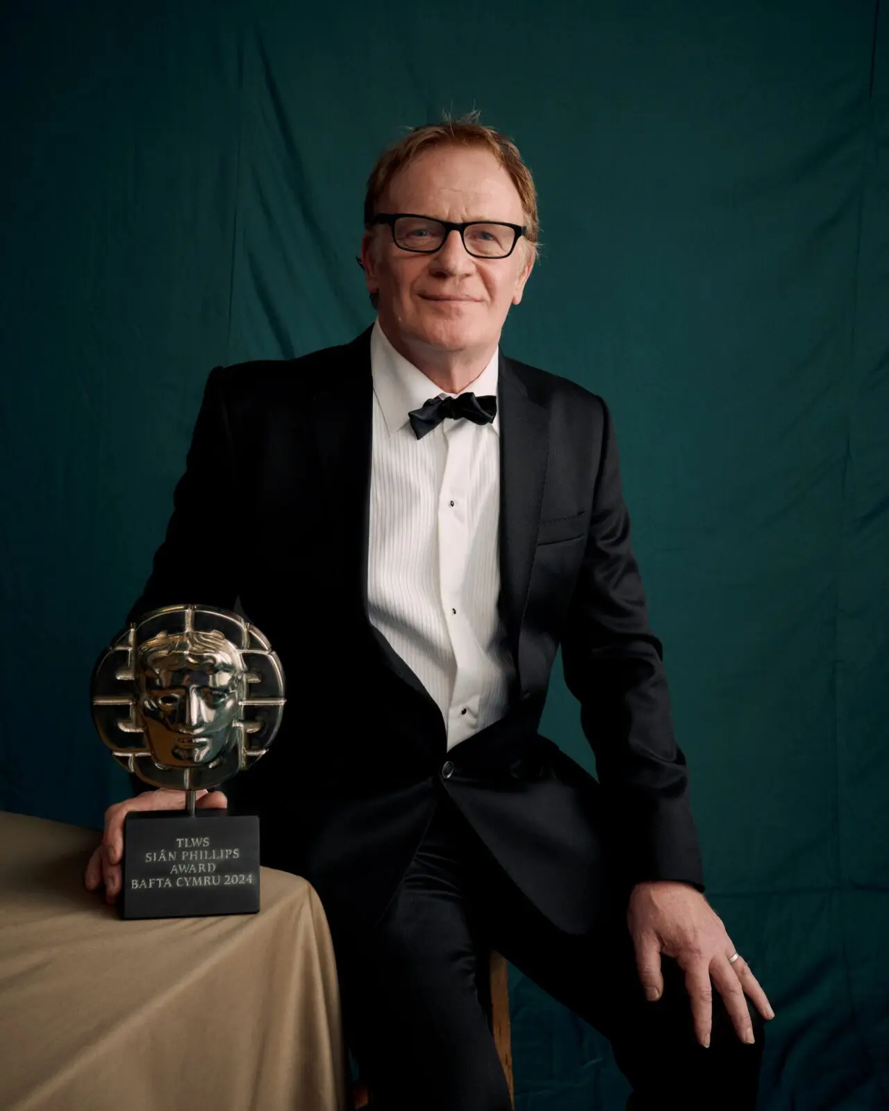 A white man in a suit with short grey hair and black-rimmed glasses. He is wearing a black tuxedo and bow tie and posing with his BAFTA.