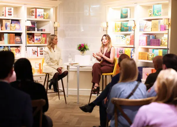 A woman in a dark red leather waistcoat and trousers sat on a high stool being interviewed by a blonde woman wearing an all white outfit.