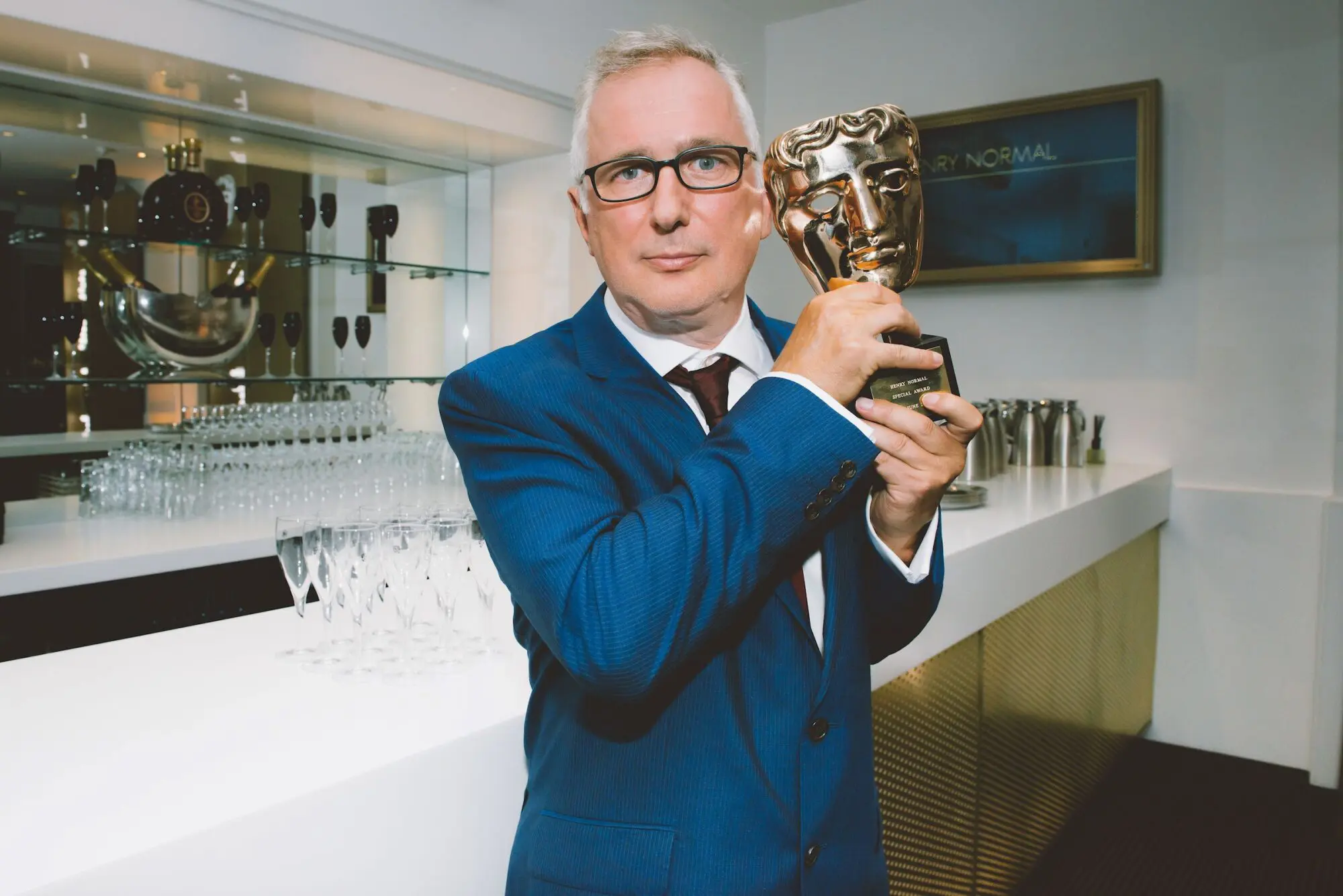 Henry Normal. A white man with greying hair, white on the sides, and black rimmed glasses. He is wearing a white shirt, black tie, blue blazer and is holding a BAFTA award.