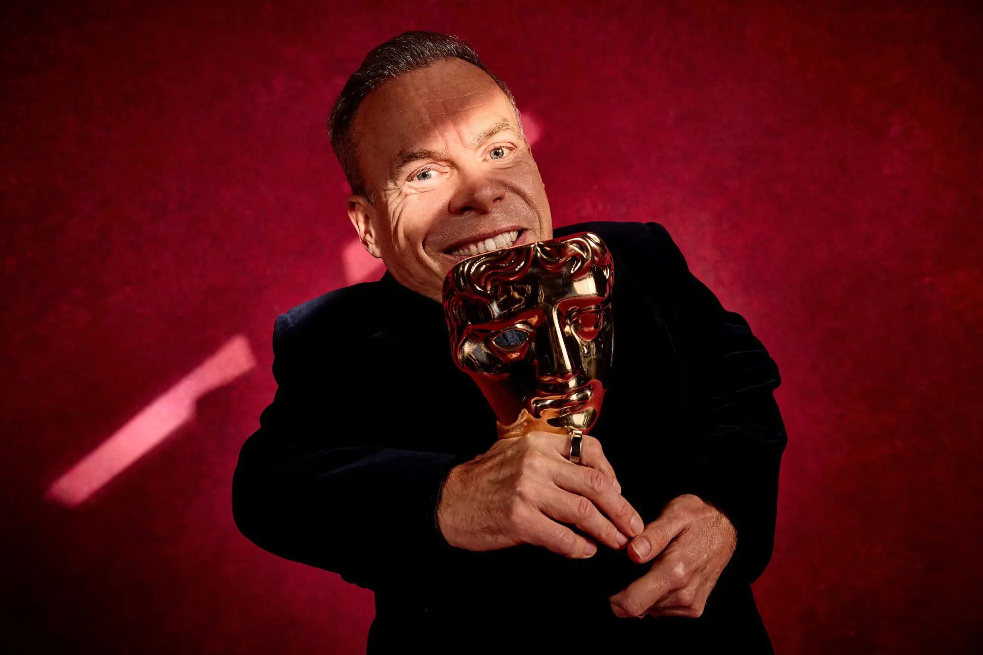 Warwick Davis. A man with thick grey hair wearing a navy velvet suit jacket and holding a BAFTA award.
