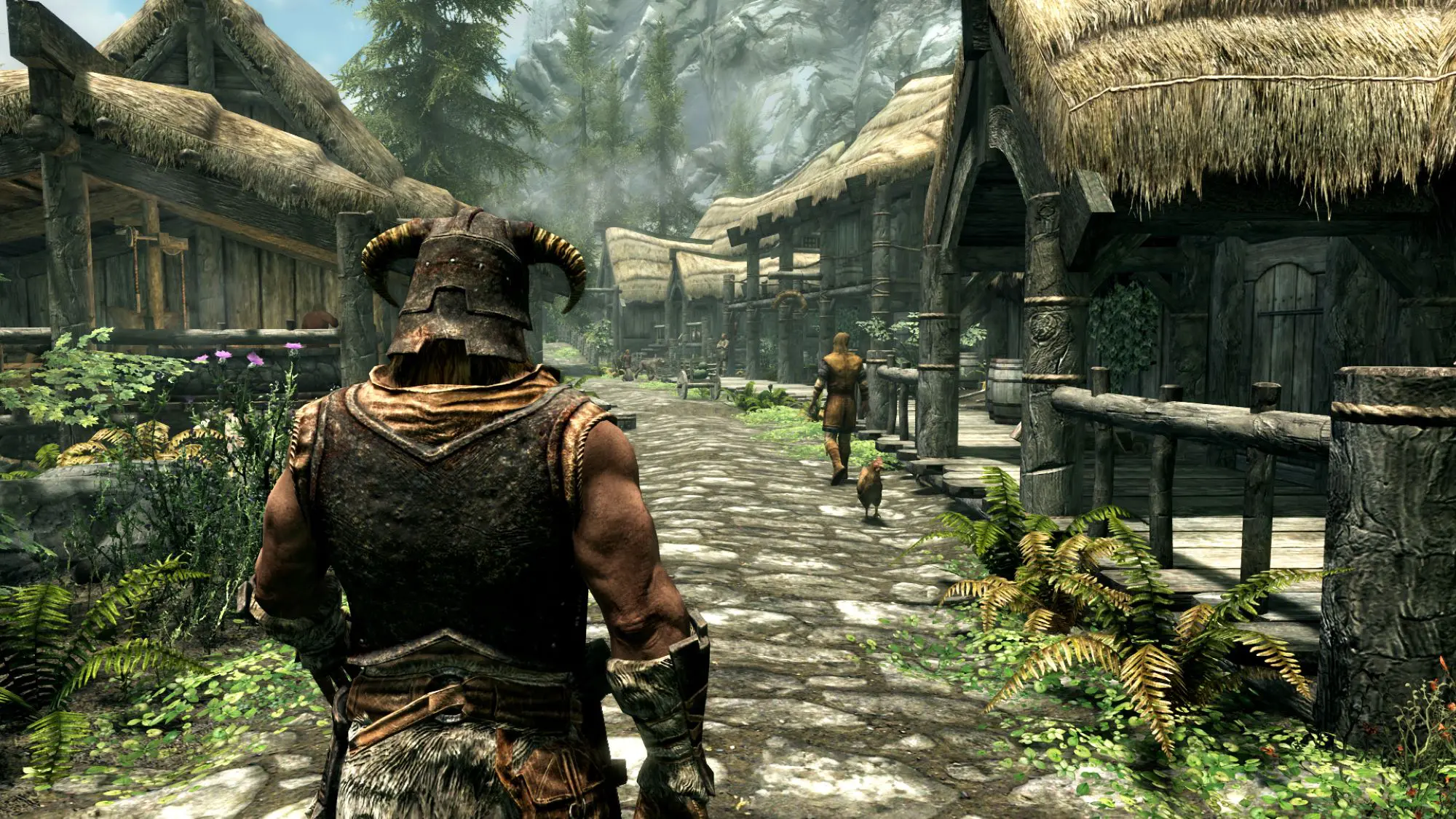 A still from The Elder Scrolls V: Skyrim game. It shows a figure in sleeveless armour and a large helmet walking into a village.
