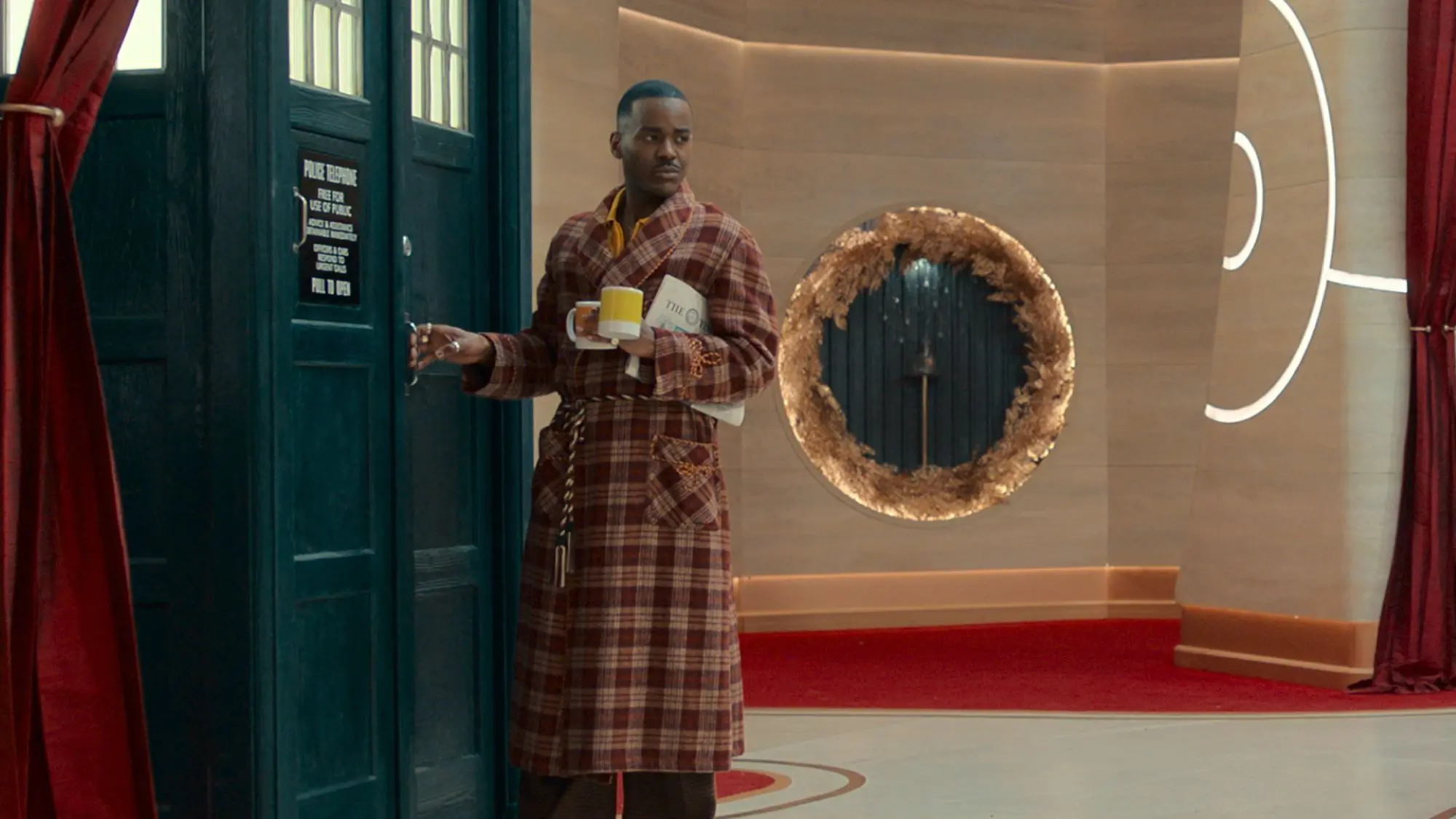 A still of Ncuti Gatwa in Doctor Who. A man with short cropped black hair wearing a tartan dressing gown and holding a yellow pantone mug. He is is about to enter a large old-fashioned phone box.