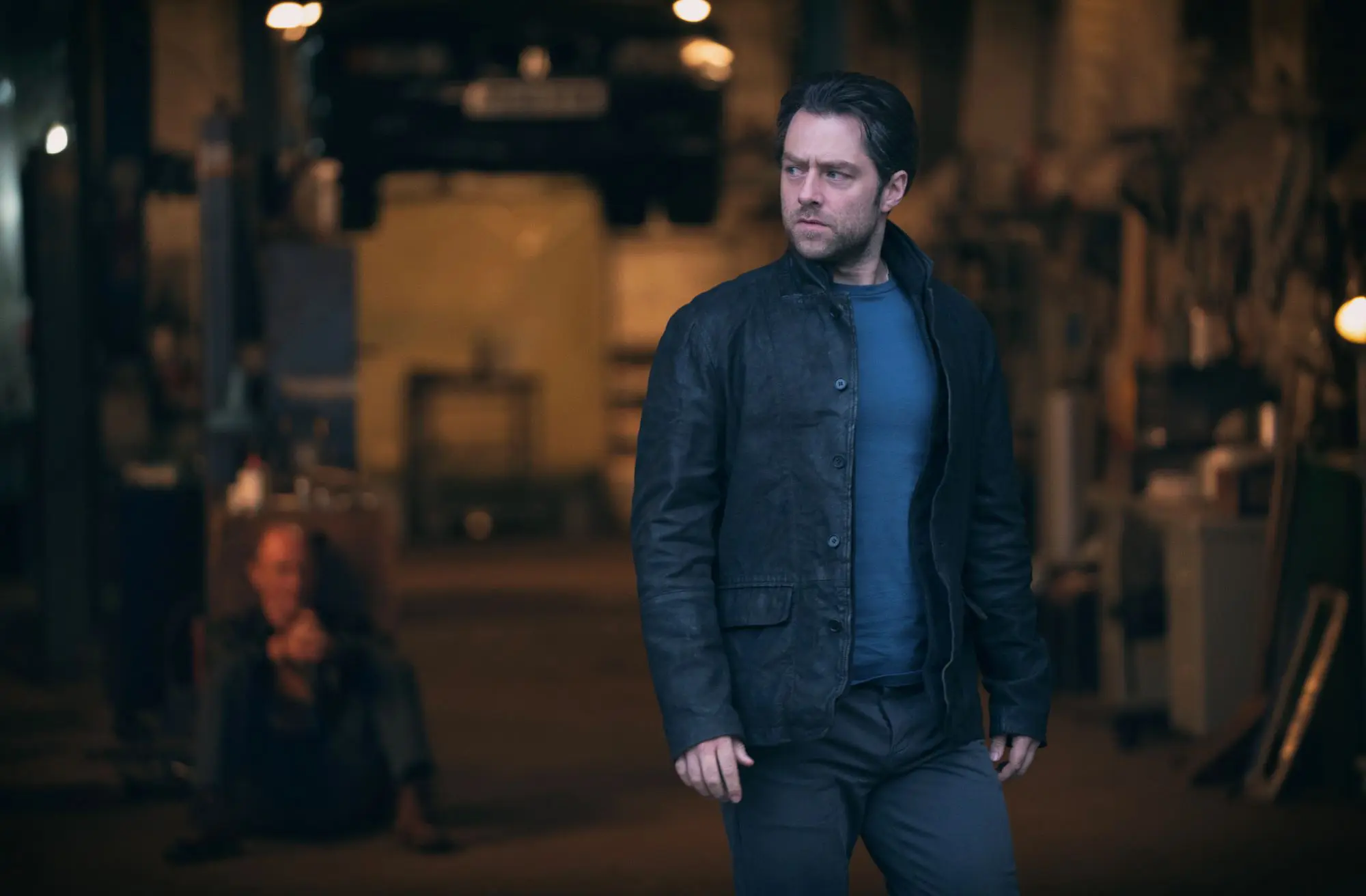 A still of Richard Rankin in Rebus. A man with thick dark hair wearing a blue sweater and dark jacket.
